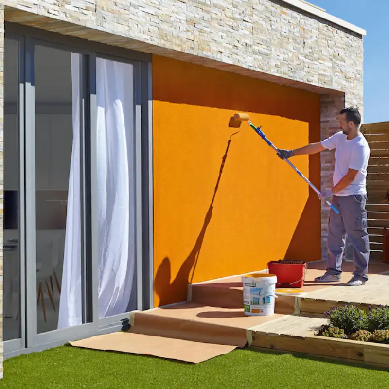 Hombre aplicando pintura o recubrimiento naranja sobre la fachada de una casa o edificio usando un rodillo de extensión. El recubrimiento especializado detiene la humedad y es ideal para edificios corporativos en Polanco o Santa Fe.