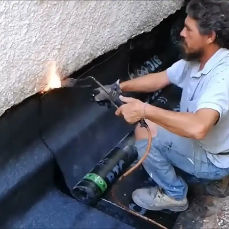 Obrero utilizando un soplete para fusionar una membrana bituminosa sobre el muro de una cimentación. Este proceso de alto espesor protege la base estructural y aísla sótanos contra la humedad ascendente en proyectos de gran escala en el Estado de México.