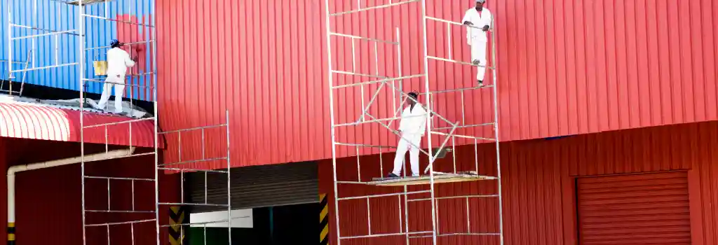 Pintores de Quantumef S.A. de C.V. trabajando en andamios en la fachada de una nave industrial roja, ilustrando el servicio de pintura profesional con 30 años de experiencia, especializada en pintura industrial en Vallejo y fachadas de edificios corporativos.